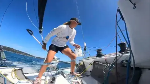 Meg Niblett A wide angle photo shows a woman stood in the middle of a sailing yacht. She is wearing a white top, black shorts and a black baseball hat. She is leaning forwards as if to compensate for the motion of the yacht.