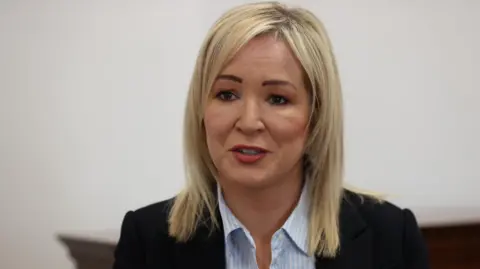 PA Media Michelle O'Neill is speaking to a camera. She has shoulder-length blond hair and is wearing a blue stripped shirt with a black blazer. 