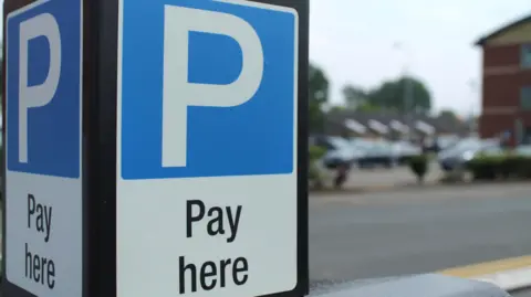 There is a large blue and white parking sign with black lettering underneath saying pay here. A car parking are can be seen out of focus in the background.