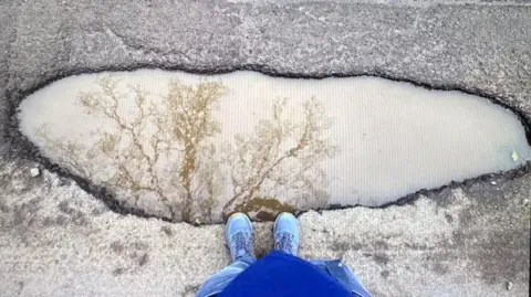 David Stone A man wearing blue jeans an blue shoes stands on the edge of a large pothole, filled with muddy water. You can see the reflection of the tree in it. 