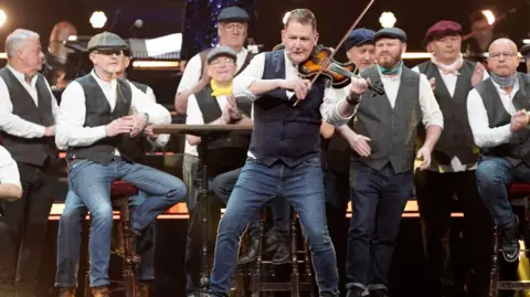Mike McGrother performs on the violin on stage backed by his choir who are standing behind him with their traditional uniform of waistcoat, white shirts and flat caps.