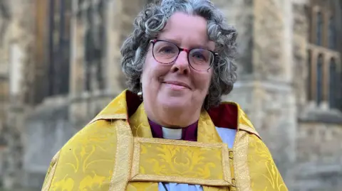 BBC Reverend Canon Mary Gregory smiling at the camera wearing black framed glasses and a yellow robe.