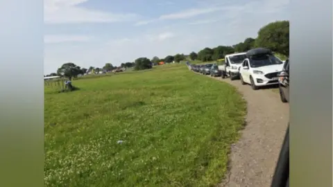 David Cars are queuing up on a road in the middle of a field.