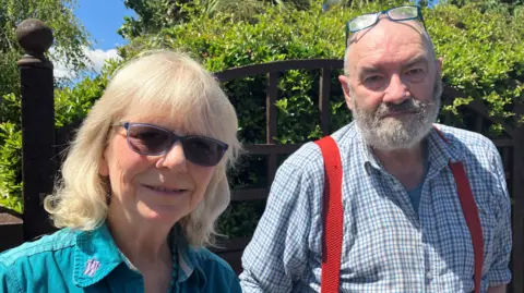 A women in turquoise top with sunglasses on. She is smiling at the camera. There is a bald man with a beard with glasses on his head standing next to her. He has a grey beard and is wearing a blue check shirt. He is looking at the camera and smiling too. 