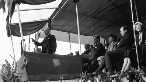Getty Images Harold Macmillan stands in front of a microphone speaking out to a crowd. One hand is held out in front of him as he addresses the crowd. People sit on seats behind him.