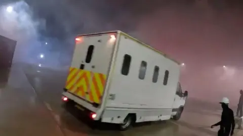 A screenshot from a video uploaded by Palestine Action. It shows a white van with curved rectangular windows and yellow and orange hi-vis decals on the back. It is night time and there is a lot of smoke in the air which is being lit up by the van's red brake lights. On the right there is a figure wearing all black and a white hard hat.