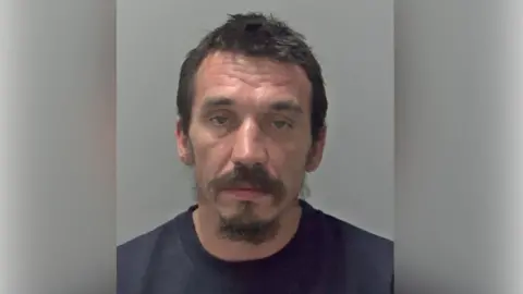 Cambridgeshire Police Fred Lamb with short dark hair and beard wearing a dark top in a police custody photo.