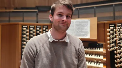 A man in a grey jumper is sitting in front of an organ, smiling at the camera. 