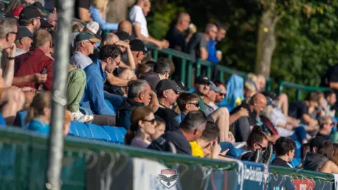 Hannah McHugh/FC Isle of Man Men and women sit in the stands of a football ground, wearing sunglasses and hats, and look towards the pitch. 