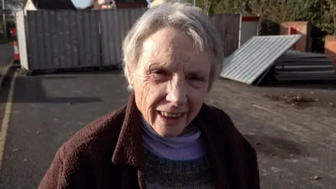 Sue Wilson, wearing a purple roll neck and brown jackets stands in front of the roadworks on Seaholme Road, Mablethorpe. She has grey hair. Metal fencing and road cones are visible in the background.