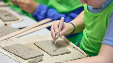 A boy wearing a light blue polo-style school shirt, uses a tool to impress lines on to a clay tablet. he is wearing a green vest over his blue top, to keep his shirt clean.