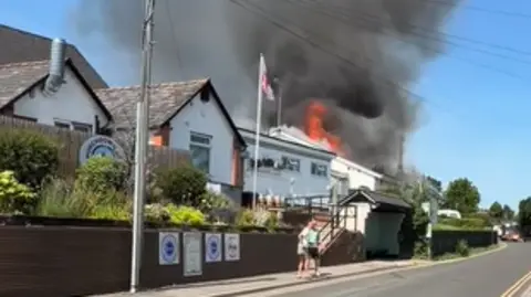 Flames and smoke billow out of a white rendered social club into the blue sky above on a village street on a summer's day. There are pedestrians standing about.