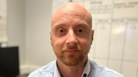 Head of patients flow Callum Allen-Ridge has a bald head, a short beard and is wearing a blue shirt.  