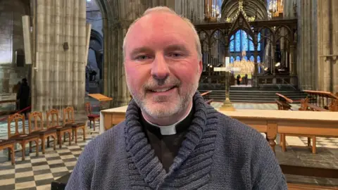 A headshot of a male reverend standing near to the front of a cathedral. He has very short grey hair, a grey beard and a grey moustache. He is smiling into the camera.