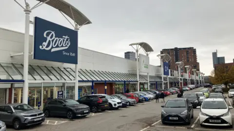 Declan Carey/LDRS A full car park with a grey coloured, long retail park building, featuring a blue Boots sign. Tall flats are in the background.
