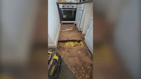 A narrow galley kitchen with an oven and white kitchen cupboards. The floor is bare boards and there is a large hole in the middle where you can see insulation. There is a black tool box in the foreground.