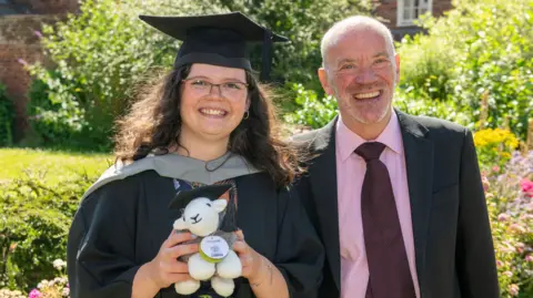 Chloe Beer and Andy Long. Beer has long wavy dark hair and wears glasses. She is wearing a cap and gown and holding a knitted sheep teddy bear with a graduation cap. Long has white shaven hair and blue eyes. He is wearing a dark suit with a pink shirt and burgundy tie. They are standing side by side and smiling at the camera. 