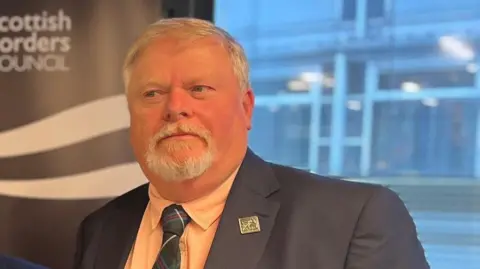 A larger man with a white goatee beard and white hair. He is wearing a blue suit and a tartan tie. He is looking slightly to the left of the camera. 