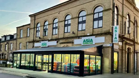 A large corner building with light brown brickwork and arched windows on the upper floor, housing a store called Asia Superstore. The ground floor features a modern glass-fronted extension displaying colourful stacks of goods inside.