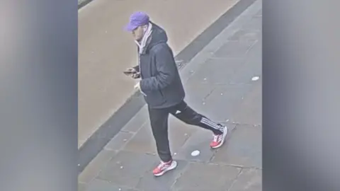 Thames Valley Police A man is walking on a section of pavement in the street. He wears a purple peaked cap, a light grey hoodie with a dark grey hooded coat over the top and holds a phone in his right hand. He is also wearing black Adidas tracksuit bottoms, which have white stripes down the sides of the legs and a paid of red trainers with white stripes on the sides.