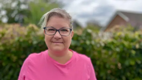 Martin Giles/BBC Sam Johnson is looking at the camera and is wearing a pink top. She has short, fair hair and is wearing spectacles. Blurred in the background are bushes and the roof of a house.