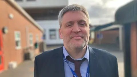 A man in blue shirt and red tie stands in front of school yard looking at camera. 