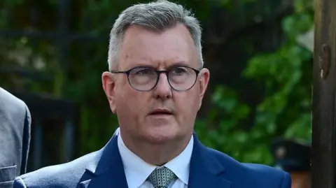 A man with grey hair and round glasses gets out of a car. He is wearing a dark grey suit, a white shirt and and green and black tie.