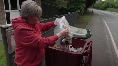 A woman in a red hoodie with blond hair is looking over an open red bin. She is holding white bin bags full of rubbish, which is falling into the bin. She is stood on the side of the road outside her house. 