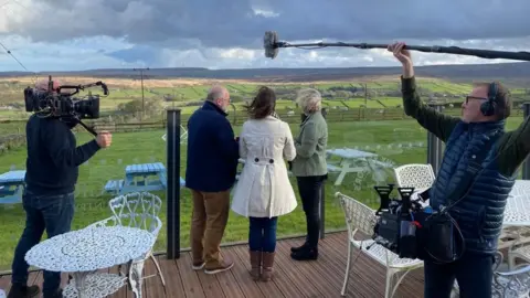 Jeanette Woodyatt Two men and a woman stand on a restaurant patio overlooking the moors of Yorkshire. Two men with cameras and a boom microphone are filming them. 