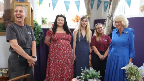 PA Media A group of five women standing in a line in the Emmaus building. On the left is Cecile Roberts a woman in a grey T-shirt smiling and looking off towards the left. On the right at the end of the row is Queen Camilla who is wearing a blue dress and looking towards the left of the frame.