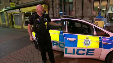 Inspector Jonathan Mellor in a police uniform getting out of a marked police car in Lincolnshire street. He has a shaved head and dark beard he is carrying his police cap in his right hand