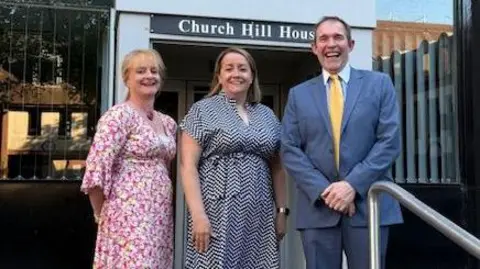 Two women and a man stood in front of a glass-fronted building called Church Hill House. All three are smiling at the camera. The woman on the left has blonde hair tied up and is wearing a patterned pink dress. The woman in the middle has shoulder length brown hair and is wearing a patterned blue dress. The man on the left has short brown hair and is wearing a blue suit, shirt and yellow tie. 