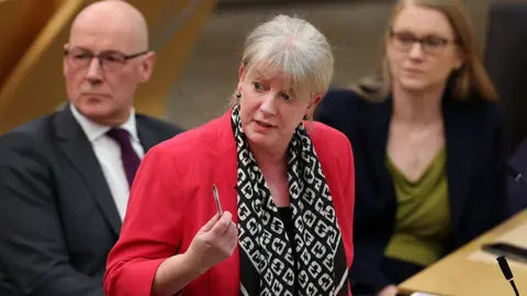 Getty Images Scottish Finance Secretary Shona Robison - a middle-aged woman with blonde hair pulled back into a ponytail, wearing a red blazer with a black and white scarf - brandishes a pen while answering a question at Holyrood. First minister John Swinney and fellow minister Shirley-Anne Somerville look on in the background, slightly out of focus