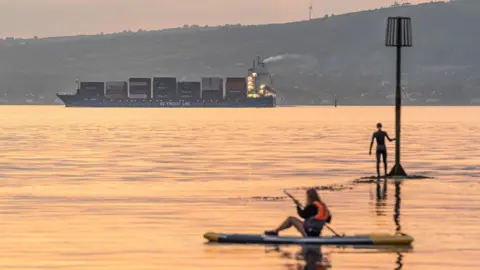 Getty Images A cargo ship  is sailing from right to left of the frame as two paddle-boarders are viewed in the foreground. It's evening and the water has an orange reflection to it. A large green hill sits behind the ship.