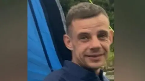 Rikki Berry, 36, who has light brown hair and wears a blue shirt, smiles at the camera with a blue tent and a grassy area in the back-drop. 