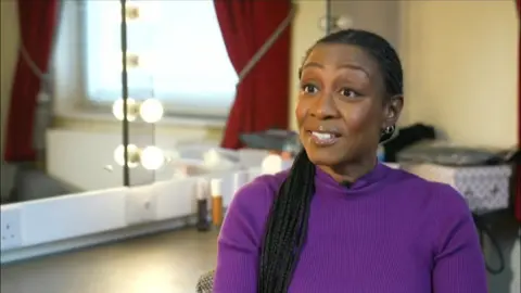 Beverley Knight, in a purple top, sitting in a theatre dressing room and looking towards an interviewer. She is sitting in front of a mirror with lightbulbs places around it, and has long, thin plaits in her hair.