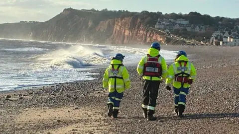 A picture of three people dressed in hi visibility clothing and blue hard hats walking on a stone beach. They are wearing fluorescent clothing.