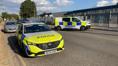 The road at an industrial estate with a police car and a police van parked up. There are metal railings and warehouses behind them, and trees dotted in the distance.