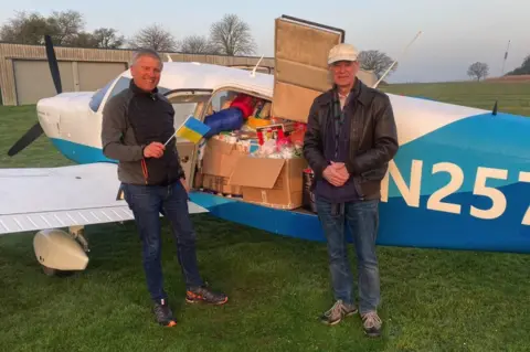 BBC Georg Gruber (left) holds a Ukraine flag and Dick Beath (right) both standing in front of the plane which shows lot of supplies loaded on it