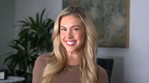 A girl with blonde hair in a brown top smiles at the camera