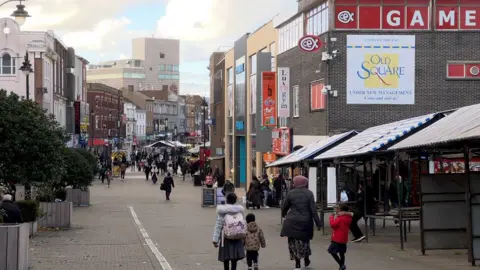 A town center with stalls, shops and people walking on the street. The stalls in the outdoor market are empty. There is a woman and three children in front of the picture, all walking down the street.