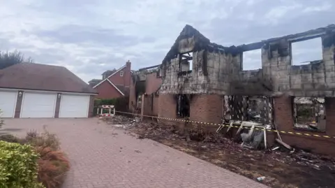 A burnt out house with yellow and black emergency service tape across it. Next to the burnt out house there is a garage with 3 white panels which has remained unscathed by the fire.