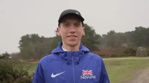 Shaun Whitmore/BBC Logan pictured in blue British running top with union flag on his left breast, smiling to camera in black cap.