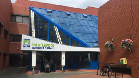 The exterior of Hartlepool Council's main building. A sign is above the front entrance. Below it, a couple pushing a pram are walking out of the doors. The building is red brick with a glass frontage.