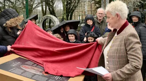 A woman with short blonde hair pulls a red cover off of a plaque. Chidren and adults in waterproof clothing watch from behind her