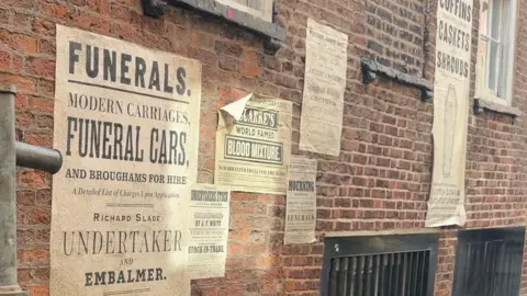 BBC News A red-brick wall covered in multiple Victorian-style posters which have been made to look rustic and messy. Most are advertising morbid services such as funerals, embalmers and a blood mixture.