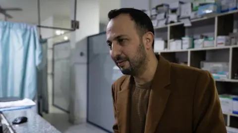 Lee Durant/BBC Doctor standing inside a hospital ward with medicine packs in the background