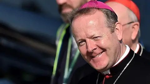 Archbishop Martin is smiling at the camera. He is wearing a pink zucchetto (Cardinal's cap) and black robes.