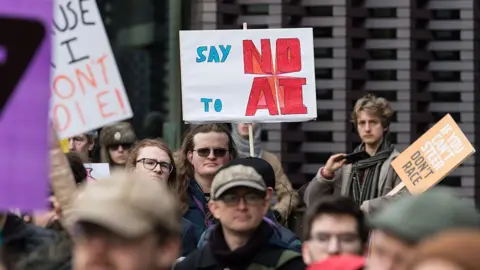 Getty Images A group of protestors photographed in a street, their faces are largely out of focus though some wearing glasses are gazing towards the camera. Centre frame is a large placard being held by one protestor, which reads "SAY NO TO AI", while a smaller placard reads "IF YOU CAN'T STEER, DON'T RACE"
