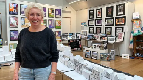 Clare Worden/BBC Lynne Burdon smiles at the camera, wearing blue jeans and a black jumper. She is surrounded by paper models of homes and other paintings. 
